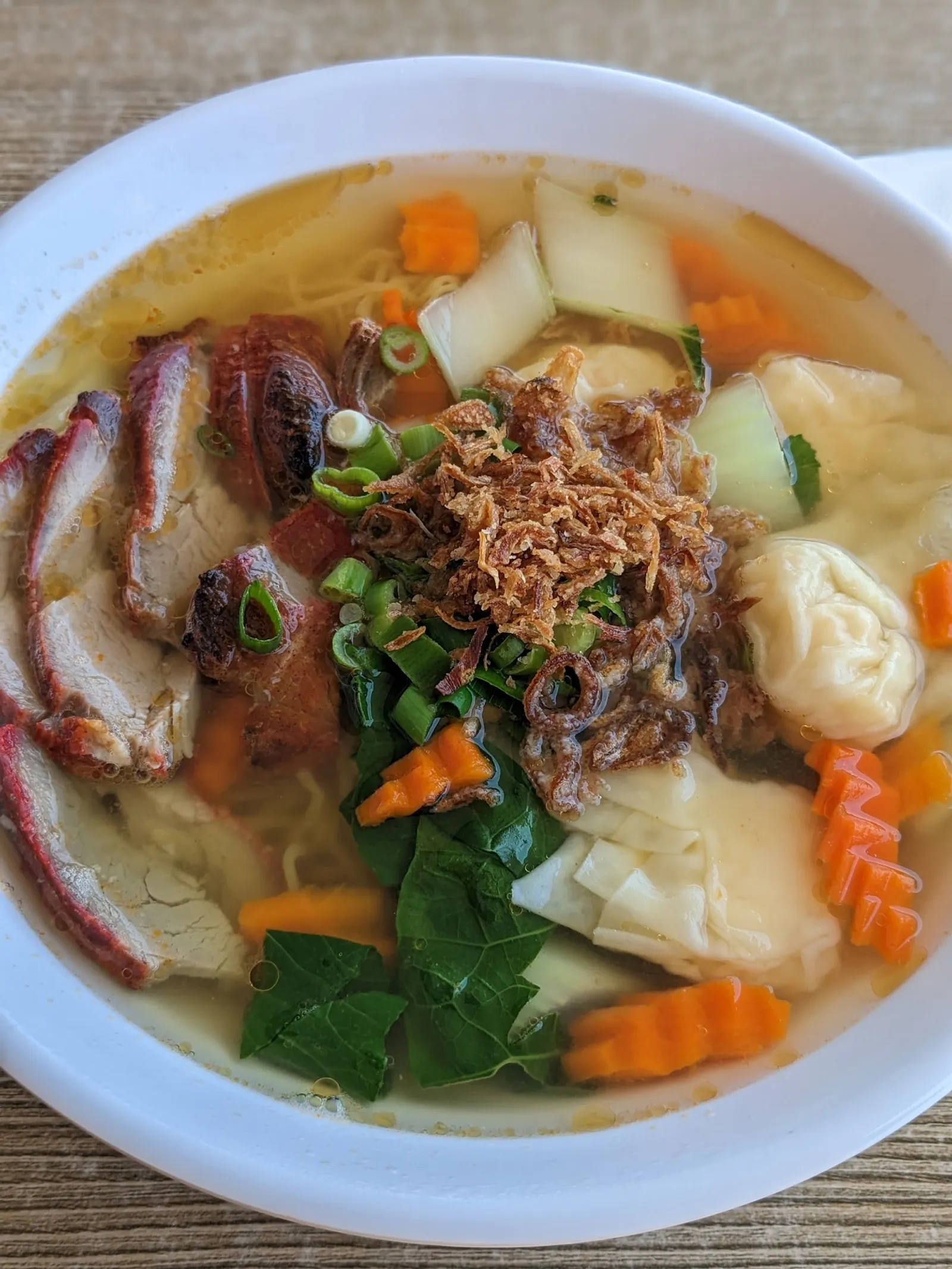 Wonton and BBQ pork noodle soup in a white bowl with char siu, wontons, and fresh herbs